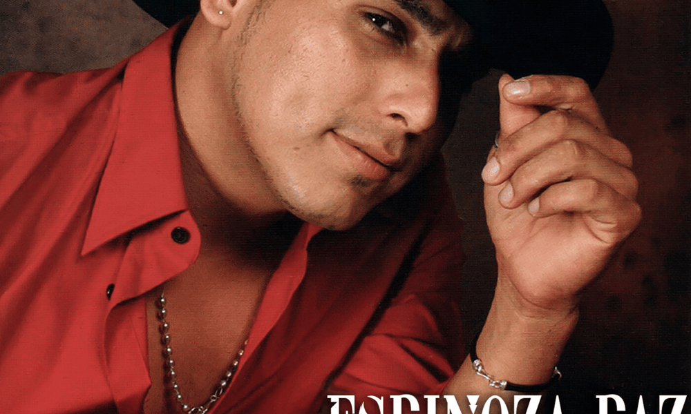 Fotografía de un hombre de sombrero negro y camisa roja, posando con la mano sobre el ala del sombrero.