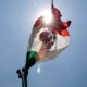 Bandera de México ondeando al sol con el escudo nacional iluminado por la luz desde atrás.