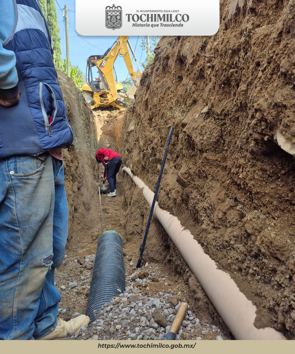 Trabajadores dentro de una zanja profunda reemplazando tubería sanitaria mientras una retroexcavadora realiza trabajos de apoyo en la calle Matamoros, Tochimilco.