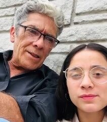 Hombre adulto de unos 50 años y una joven de alrededor de 20 grabando un video juntos frente a una pared de piedra.