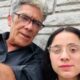 Hombre adulto de unos 50 años y una joven de alrededor de 20 grabando un video juntos frente a una pared de piedra.