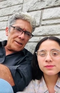 Hombre adulto de unos 50 años y una joven de alrededor de 20 grabando un video juntos frente a una pared de piedra.