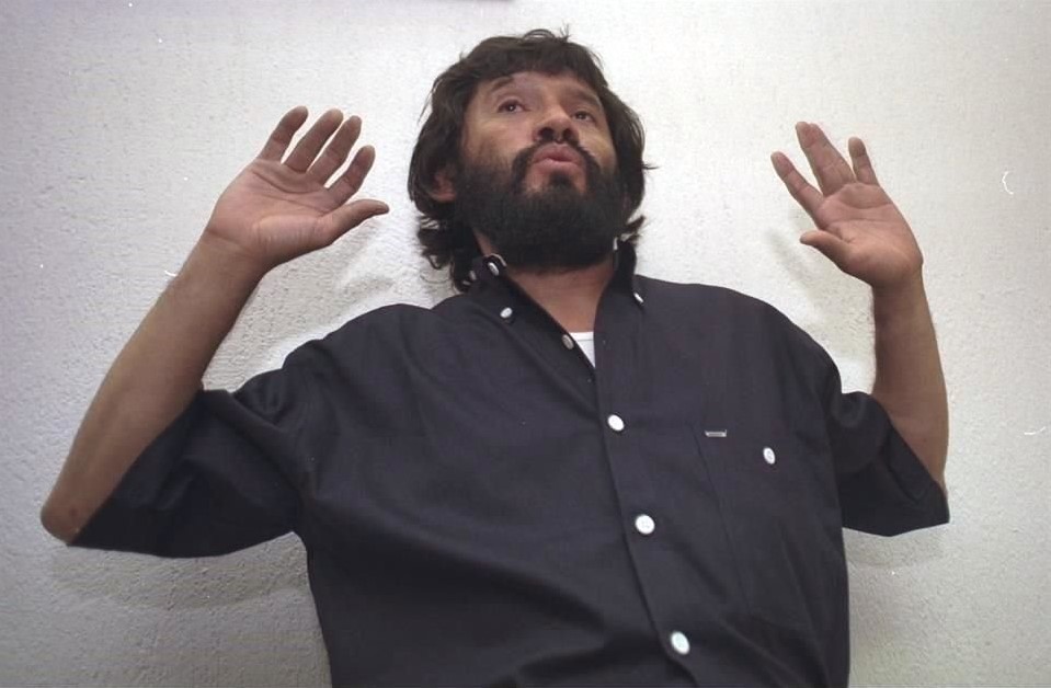 Persona con barba levantando las manos frente a un fondo blanco.