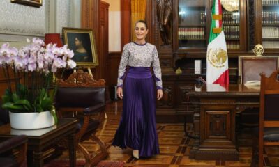 Persona caminando en un salón oficial con decoración tradicional mexicana y bandera nacional al fondo.