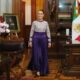 Persona caminando en un salón oficial con decoración tradicional mexicana y bandera nacional al fondo.