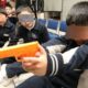 Niños sentados en un aula sosteniendo juguetes con forma de pistola, con los rostros deliberadamente cubiertos para proteger su identidad.