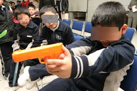 Niños sentados en un aula sosteniendo juguetes con forma de pistola, con los rostros deliberadamente cubiertos para proteger su identidad.