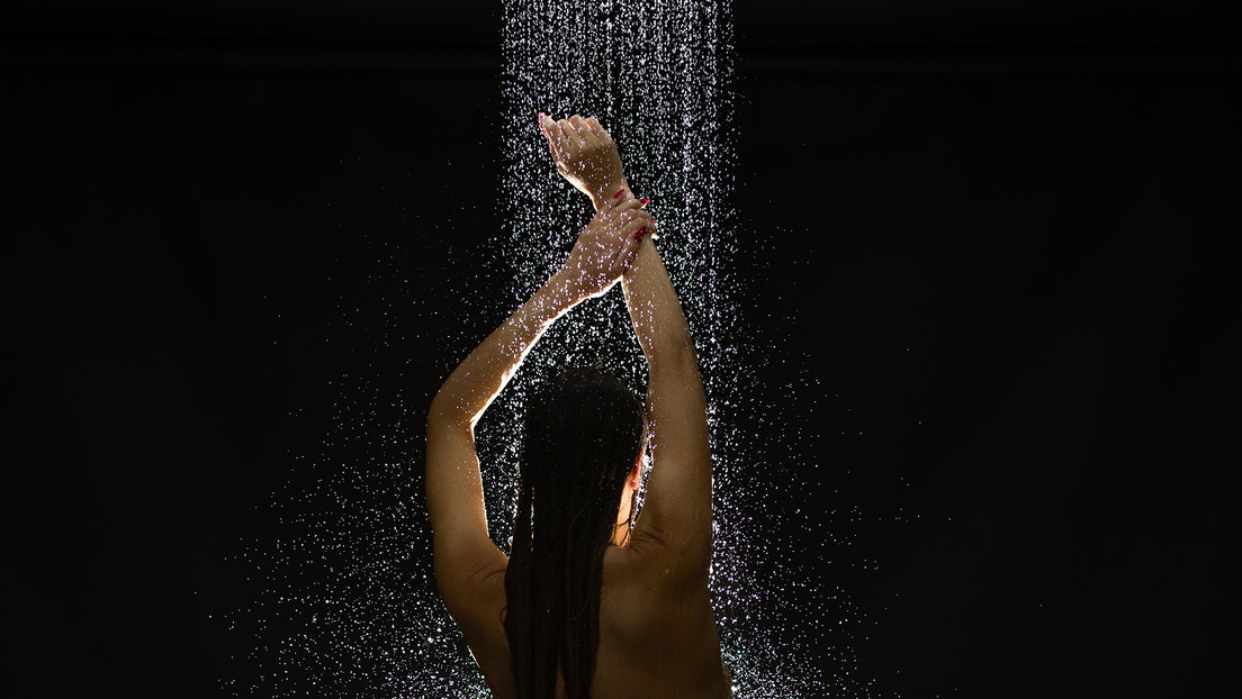 Persona bajo un chorro de agua en una ducha oscura, iluminada desde arriba.