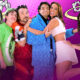 Grupo de personas vestidas con atuendos coloridos y peluche posando juntas frente a un fondo púrpura.