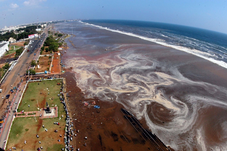 Vista aérea de una playa con manchas oscuras y sedimentos sobre el mar, muestra zonas afectadas tras un tsunami cerca de una costa urbana.