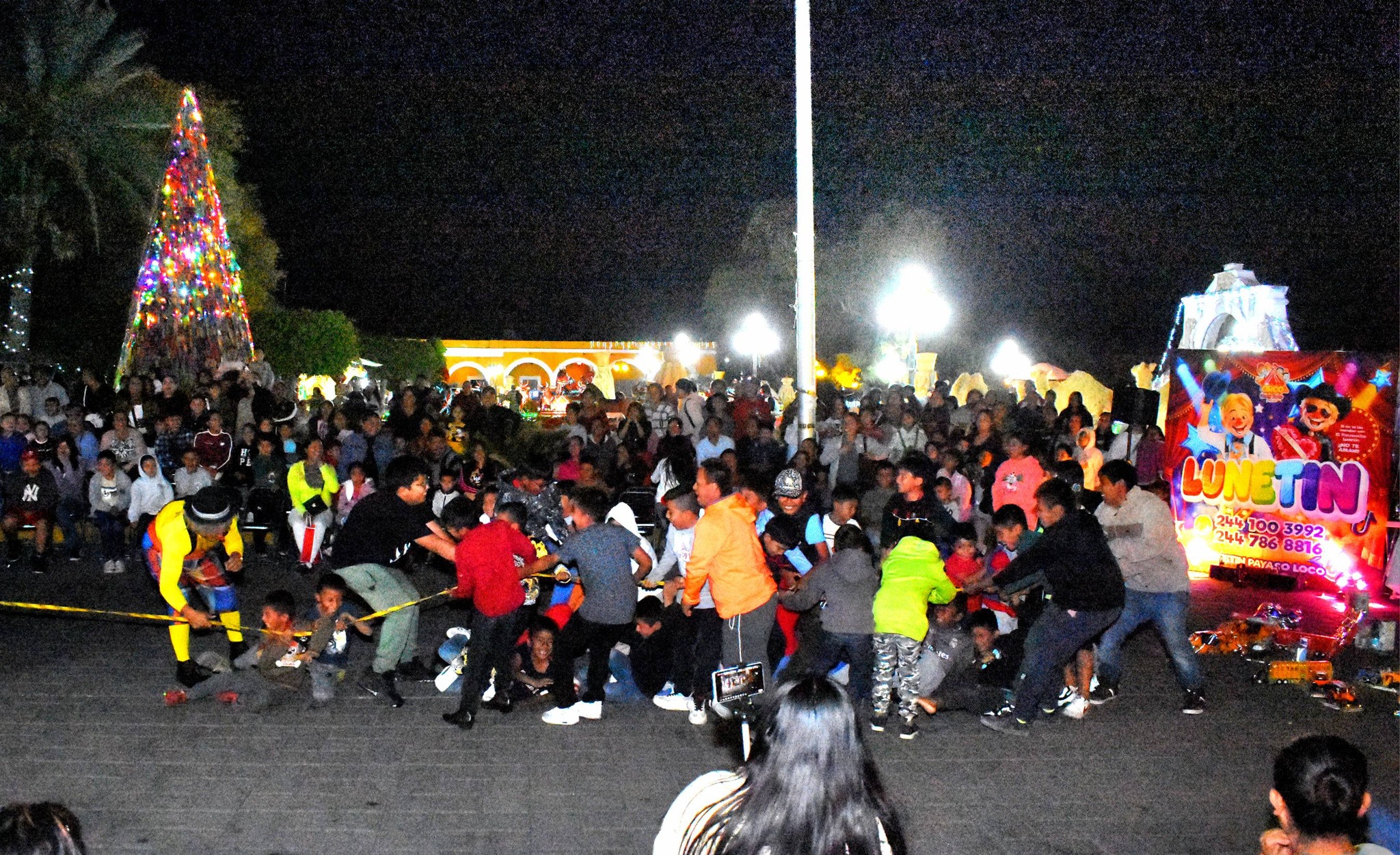 Niñas y niños participando en la entrega de juguetes durante la celebración del Día de Reyes en la Cabecera Municipal de Huaquechula, con familias reunidas de noche en la plaza.