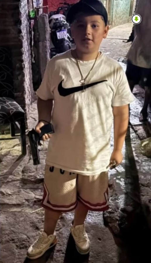 Fotografía de un niño posando con lo que parece ser un arma de fuego en la mano, en un espacio público nocturno.
