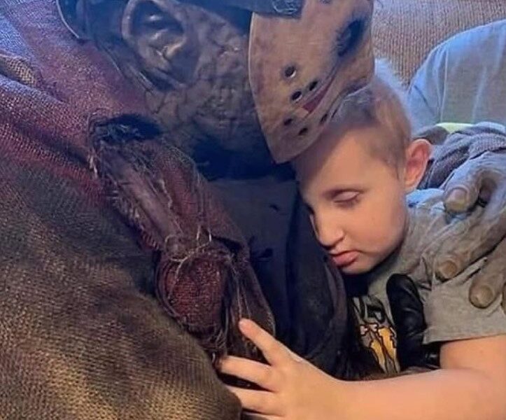 Cosplayer de Jason Voorhees acompañando de forma afectuosa a un niño pequeño, en un momento de consuelo emocional.