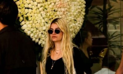 “Mujer joven con gafas oscuras y cabello rubio frente a un gran arreglo floral blanco, en un entorno tipo funeraria o acceso a recinto.”