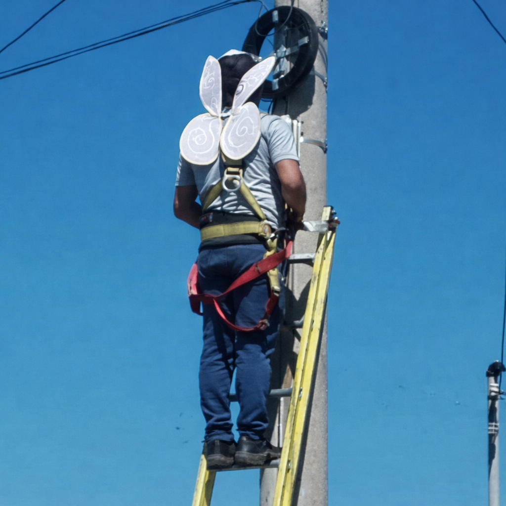 Trabajador en altura con equipo de seguridad y alas simbólicas mientras realiza labores en un poste eléctrico