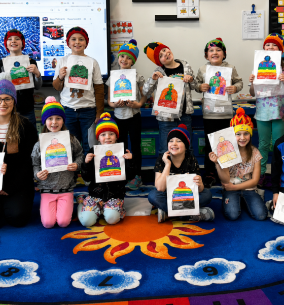 Profesora y alumnos muestran gorros tejidos inspirados en dibujos realizados por los estudiantes dentro de un salón de clases.