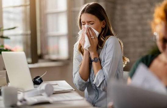 Mujer con síntomas de alergia cubre su nariz con un pañuelo mientras trabaja en una oficina, imagen representativa del Día Mundial de la Alergia y la importancia del diagnóstico oportuno.